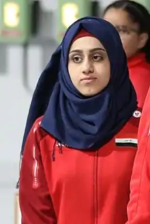 Fatimah Abbas is seen wearing a black hijab, red track suit jacket, and black athletic pants.