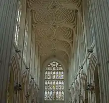 Interior of large building with a stained glass window at the far end. Above is a fan vault ceiling and on either side rows of arches.