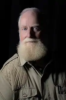 Action portrait of a man in his fifties with a bushy, strawberry-blond beard seated while speaking wearing a safari jacket and gold watch