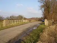 road with concrete pillars and white painted handrails where the drainage ditch passes underneath. A stand of bare trees on the right and some bare hedgerows on the left show this is winter