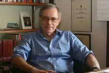 A grey-haired bespectacled man wearing a light blue shirt and sitting on a chair behind a desk; behind him is a bookshelf and a wall mounted with certificates and awards