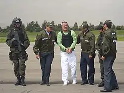 A handcuffed man in a light green T-shirt, flak jacket and white pants being escorted by two men on either side in green jackets and caps with the Spanish word "Policia" identifying them as police officers. To the left is a soldier in body armor and helmet, carrying an assault rifle. At the right is another police officer, his back to the camera, slightly obscuring another similarly equipped soldier.