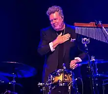 John Robinson standing behind the drums, wearing a black suit, hand on his heart to acknowledge applause