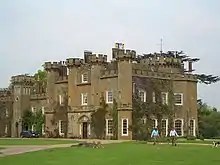 A battlemented stone building with two people, a dog, and a car in front.