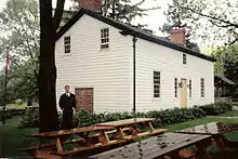A white house in three-quarter perspective. In the foreground are several picnic benches. To the left is a tall tree with a man standing beside it. To the right, partly obsured by the house, is an old well.
