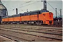Three ALCO FA locomotives parked on a track in the yard. A railroad worker is standing near the lead locomotive.