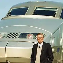 Cropped colour photograph of man in dark jacket, shirt and tie standing in front of French TGV high-speed train.